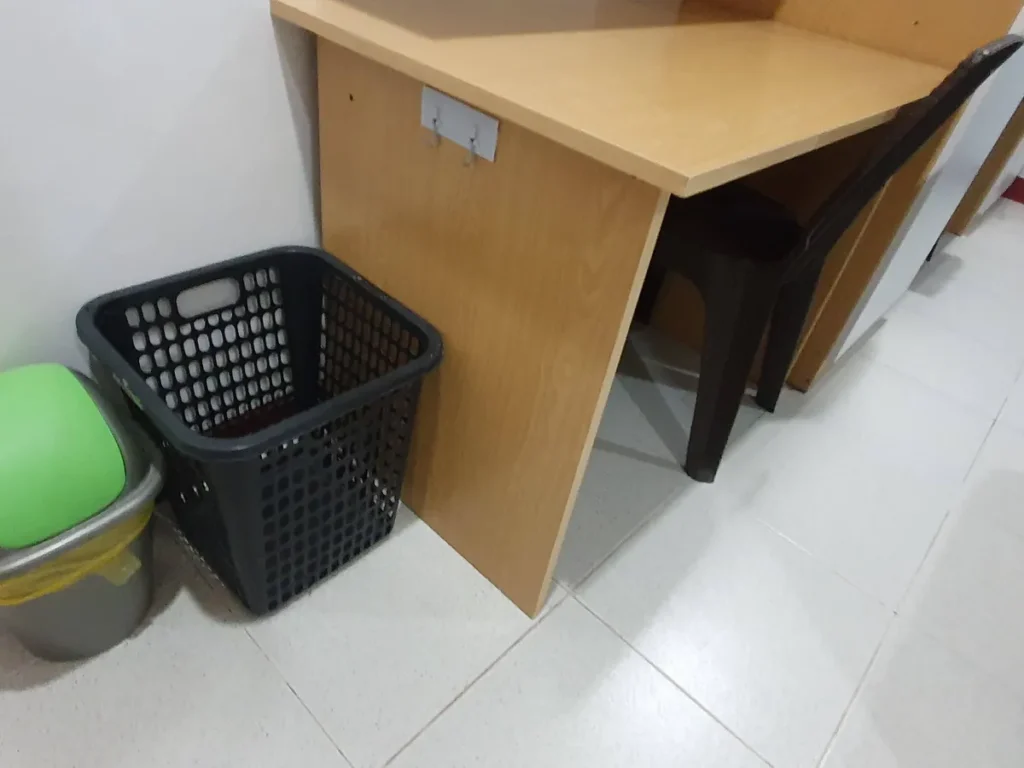 필리핀 세부 CG 바닐라드 어학원 기숙사 2인실로, 책상 다리 옆에 검정 빨래바구니와 초록 뚜껑 쓰레기통이 나란히 놓인 수납 공간 디테일입니다.