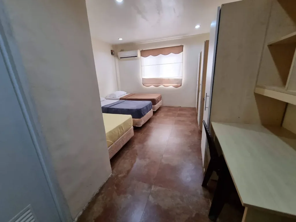 필리핀 세부 CG 바닐라드 어학원 기숙사 3인실로, 노랑·남색·갈색 침구의 싱글 침대 3개와 갈색 물결무늬 커튼 창문, 에어컨이 보이는 전체 침실입니다.