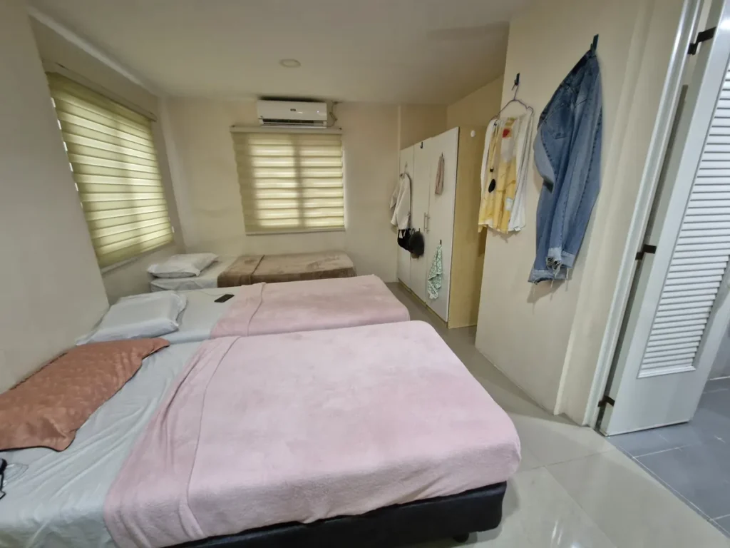 필리핀 세부 CG 바닐라드 어학원 기숙사 3인실로, 분홍색 침구 싱글 침대 여러 개와 에어컨·옷걸이·흰색 옷장이 보이는 침실입니다.