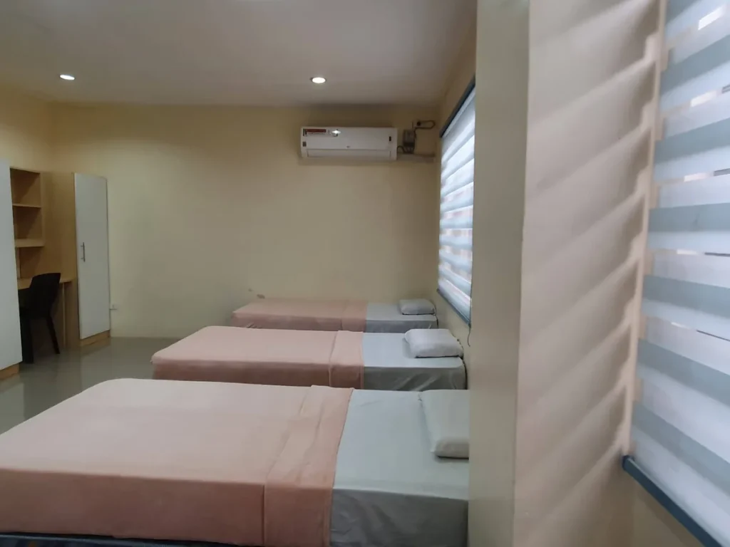 필리핀 세부 CG 바닐라드 어학원 기숙사 4인실로, 책상 겸용 옷장과 에어컨 아래 나란히 놓인 3개의 창가 침대가 보이는 실내입니다.