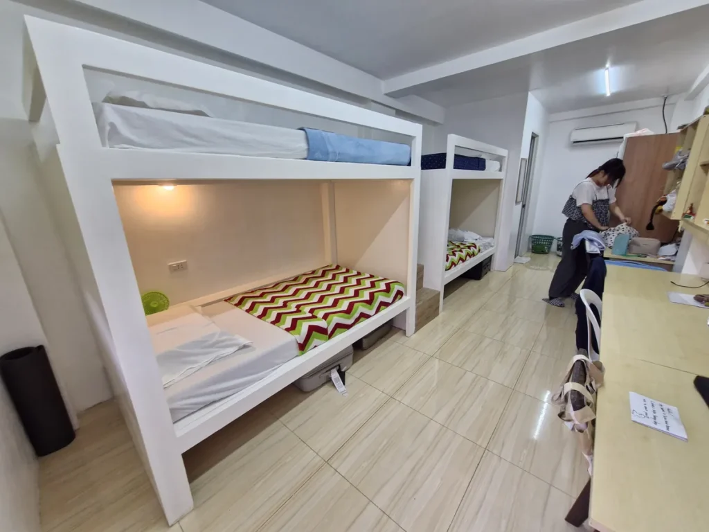 필리핀 세부 CG 바닐라드 어학원 기숙사 4인실(2층침대)로, 흰색 2층 침대에 체크무늬 이불이 깔려 있고 침대 밑 수납함이 놓인 다른 동 객실입니다.