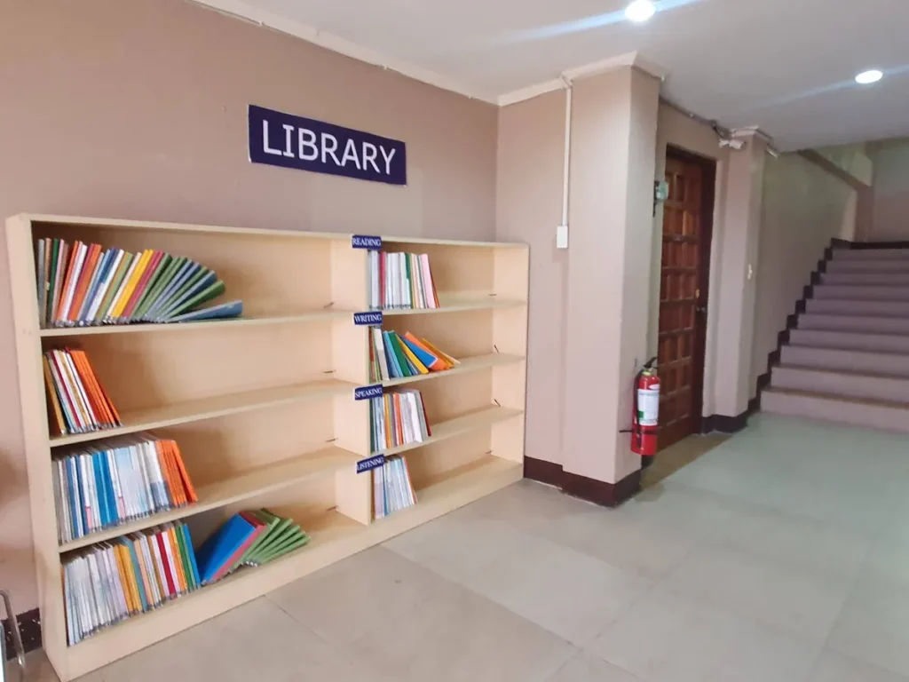 필리핀 세부 CG 바닐라드 어학원 강의실 교재로, LIBRARY 표지판 아래 4단 책장에 영역별 색깔 교재가 꽂혀 있고 옆에 계단이 보입니다.