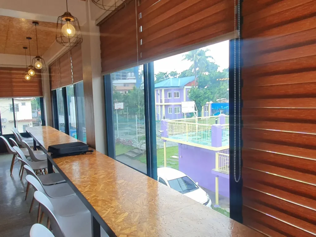필리핀 세부 CG 바닐라드 어학원 매점으로, 통유리창 앞 긴 바 테이블에 서류가방이 놓여 있고 창밖으로 야자수와 주택가가 보이는 창가 좌석입니다.