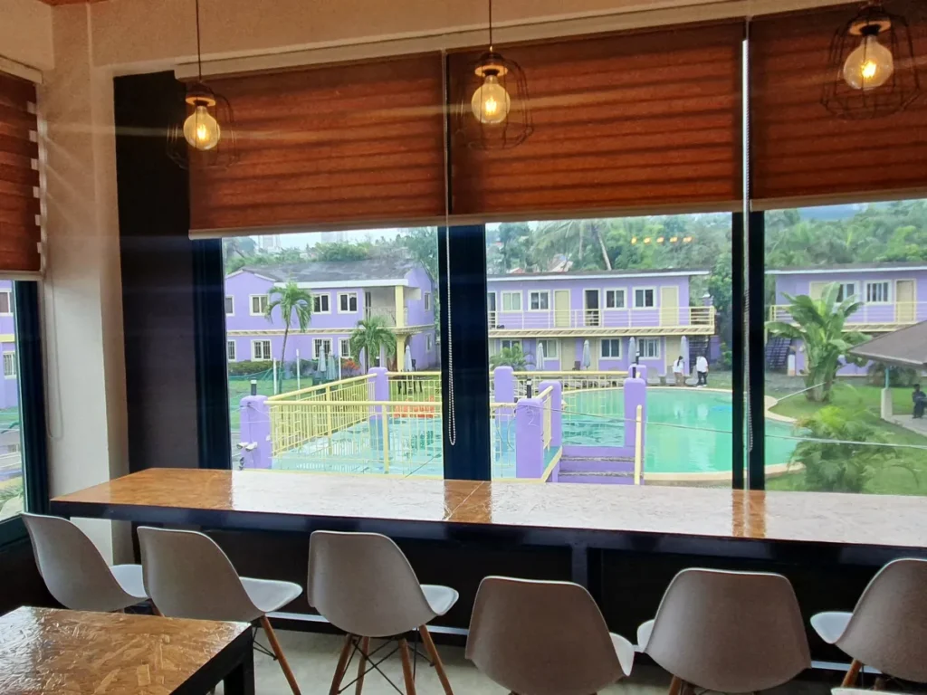 필리핀 세부 CG 바닐라드 어학원 매점으로, 나무 블라인드 조명이 걸린 통유리창 너머로 보라색 기숙사 건물과 넓은 수영장이 펼쳐진 매점 전망입니다.