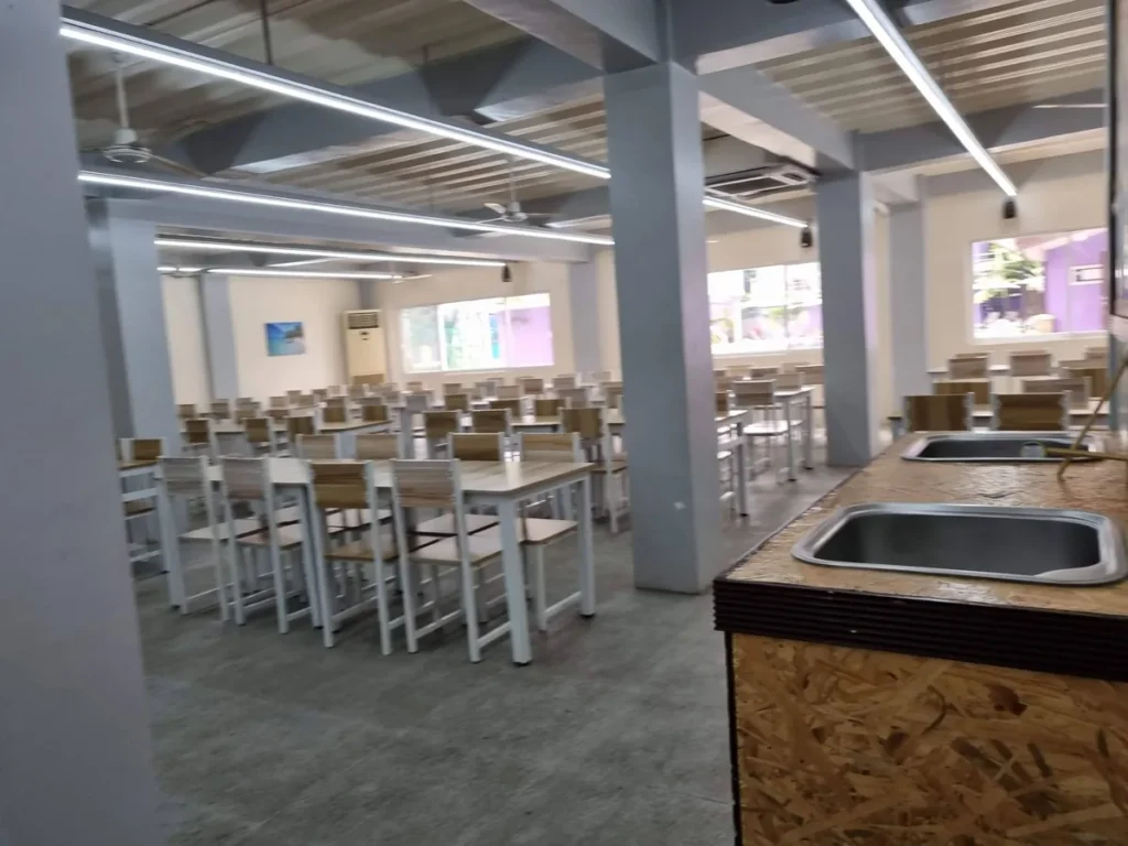 필리핀 세부 CG 바닐라드 어학원 식당으로, 목재 하부장 위 스테인리스 개수대가 설치된 조리대와 안쪽 테이블·의자가 보이는 모습입니다.
