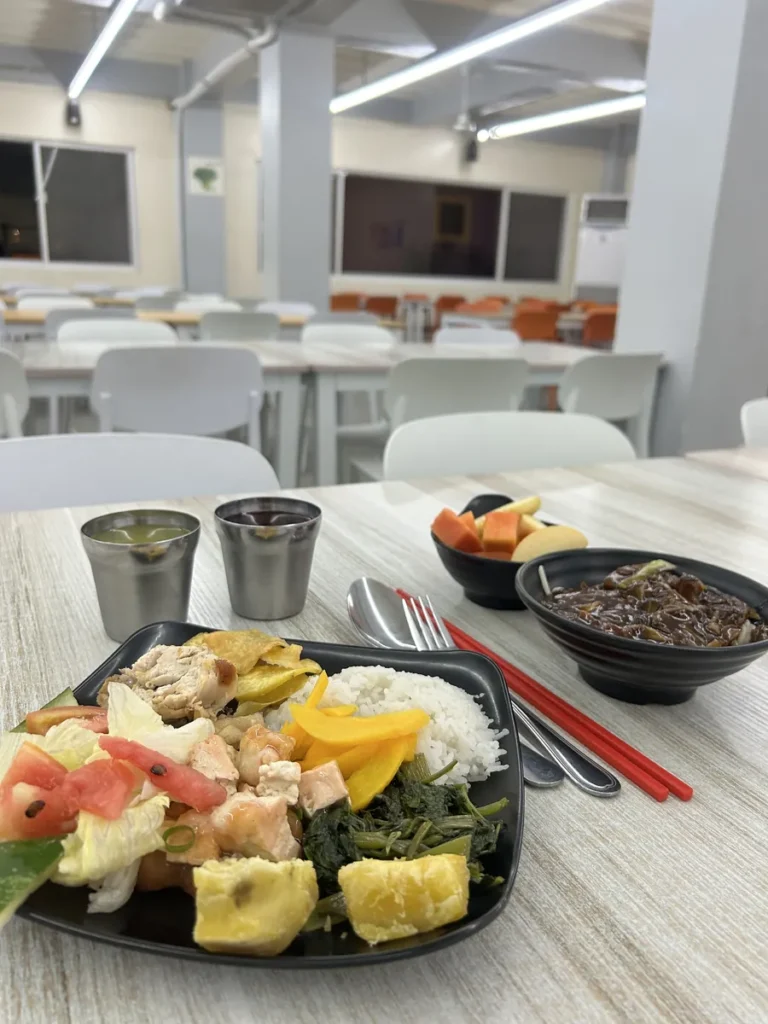 필리핀 세부 CG 바닐라드 어학원 식당 식사로, 테이블이 늘어선 넓은 홀을 배경으로 밥·나물·치킨·과일이 담긴 개인 식판이 놓여 있습니다.