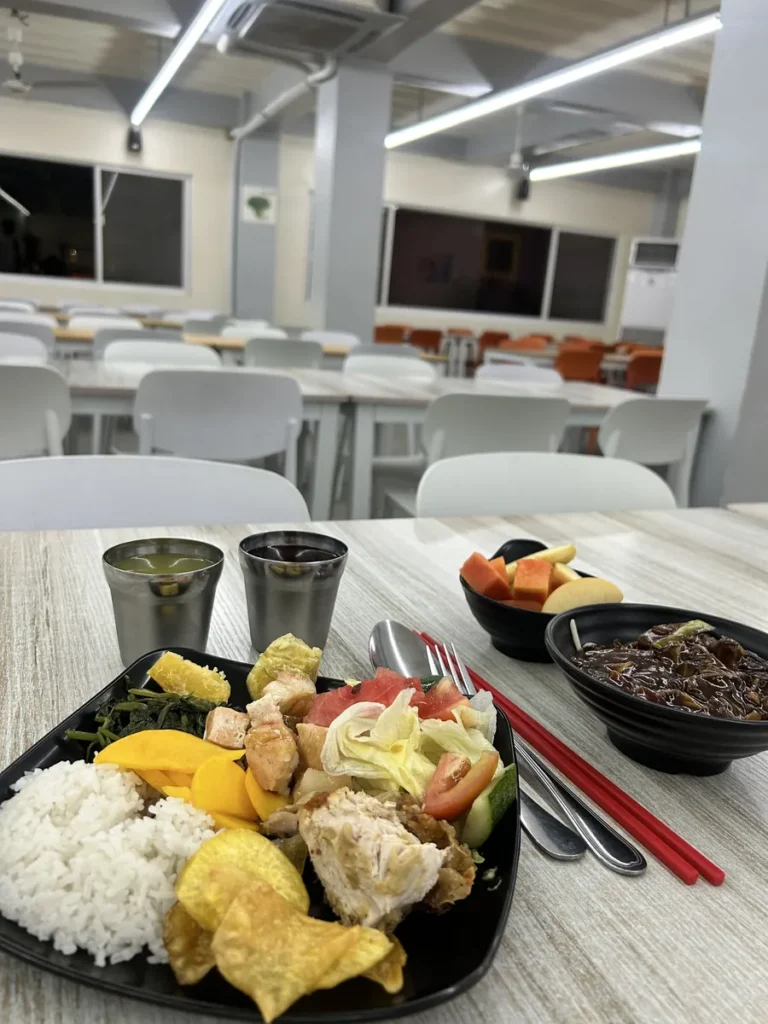 필리핀 세부 CG 바닐라드 어학원 식당 식사로, 야간 조명이 켜진 홀에서 밥·시금치나물·감자칩·과일 식판과 음료 두 잔이 놓여 있습니다.