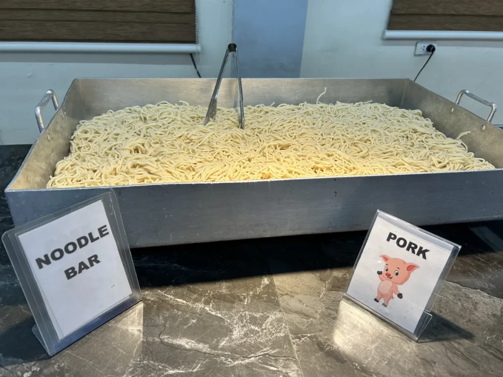 필리핀 세부 CG 바닐라드 어학원 식당 식사로, NOODLE BAR와 PORK 팻말이 세워진 조리대에 삶은 면이 큰 철제 트레이 가득 담겨 있습니다.