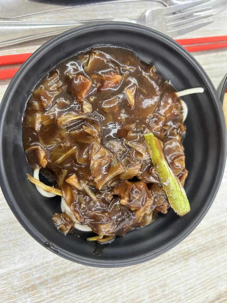 필리핀 세부 CG 바닐라드 어학원 식당 식사로, 검은 그릇에 양파와 청양고추를 곁들인 진한 갈색 소스의 조림 요리가 포크와 함께 담겨 있습니다.