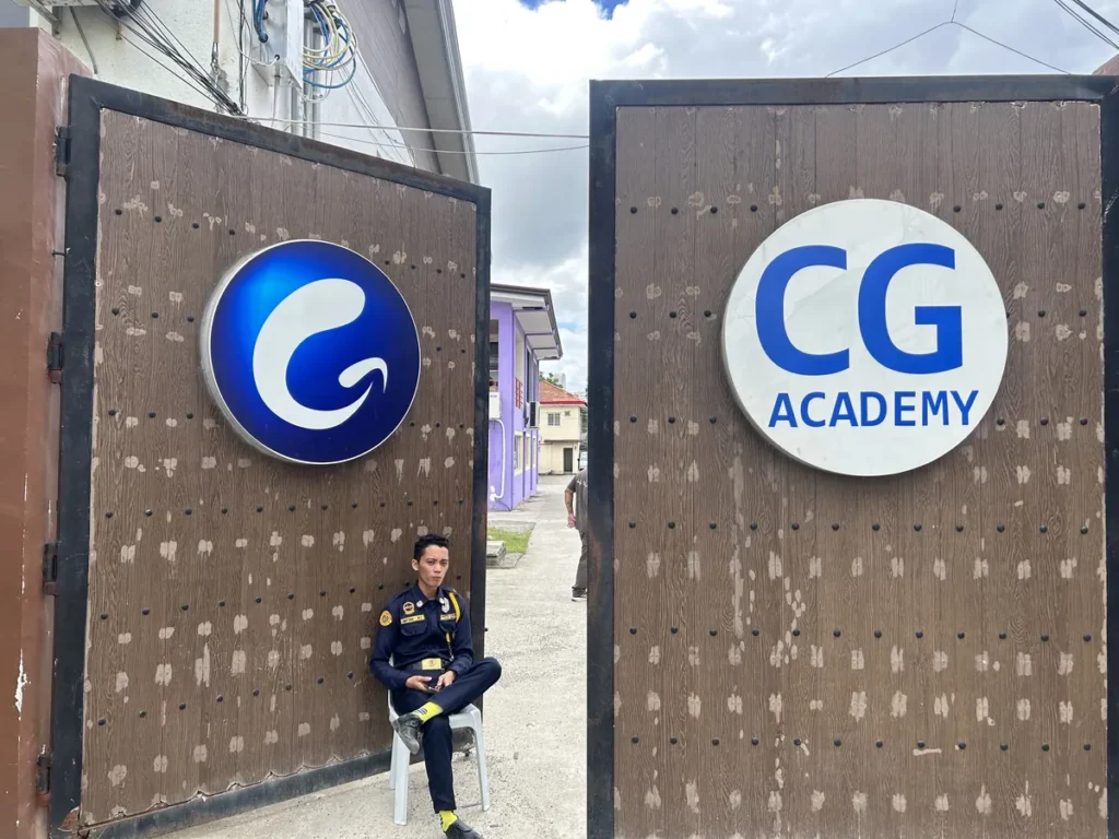 필리핀 세부 CG 바닐라드 어학원 입구로비로, 갈색 우드 게이트에 CG 로고와 CG ACADEMY 간판이 걸려 있고 경비원이 의자에 앉아있는 정문입니다.