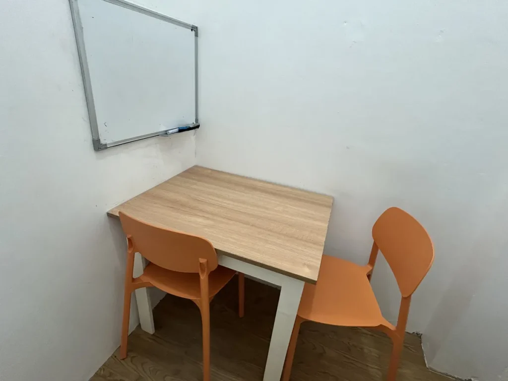 필리핀 세부 CG 스파르타 어학원 강의실 일대일 수업실로, 빈 화이트보드 아래 원목 책상과 오렌지색 의자 두 개가 마주 놓인 모습입니다.