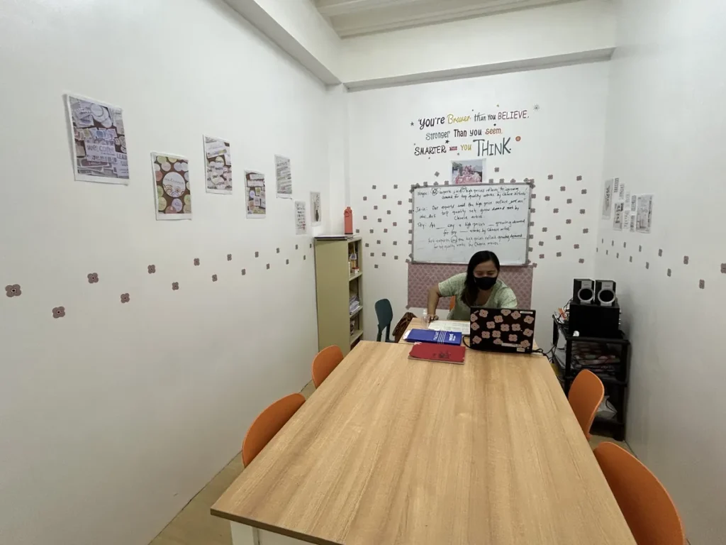 필리핀 세부 CG 스파르타 어학원 강의실 그룹반으로, 마스크를 쓴 선생님이 화이트보드 앞 책상에 앉아 있는 원목 테이블의 소규모 공간입니다.