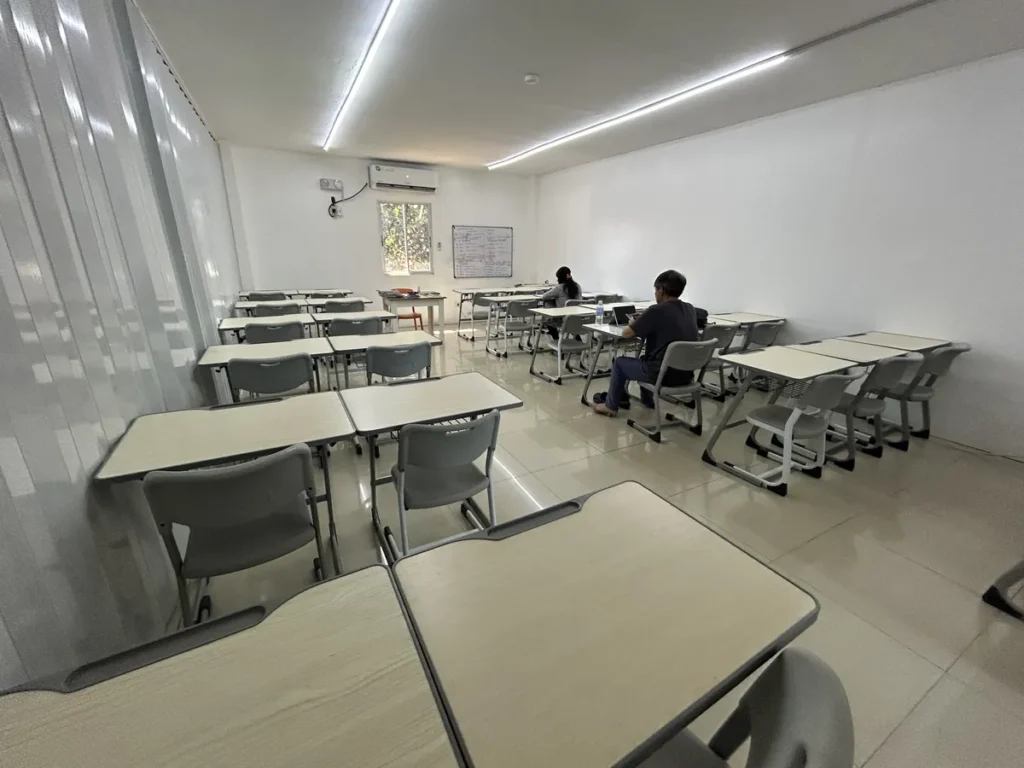 필리핀 세부 CG 스파르타 어학원 자습실로, 회색 책상과 의자가 줄지어 놓이고 에어컨과 화이트보드가 설치된 밝은 흰색 벽 학습공간입니다.