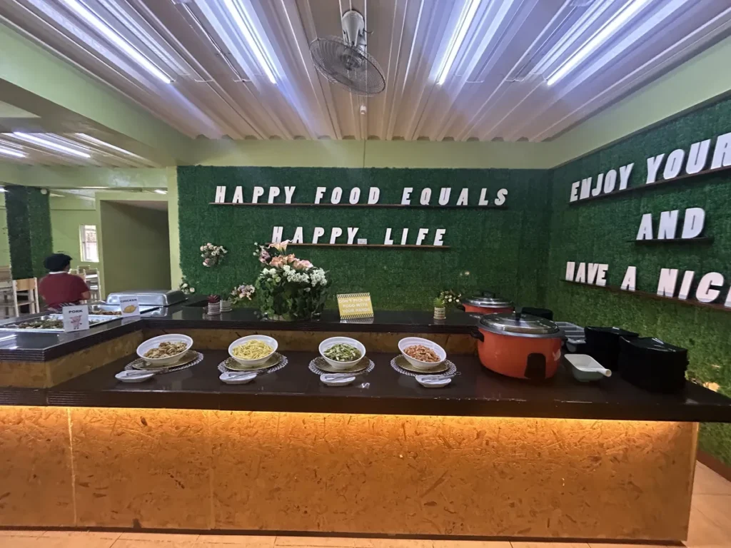 필리핀 세부 CG 스파르타 어학원 식당으로, 초록 벽 문구 아래 국수와 나물 반찬, 전기밥솥이 놓인 배식대입니다.
