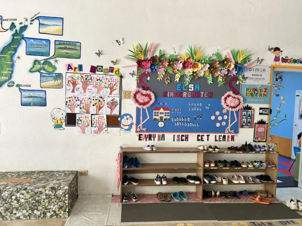 필리핀 세부 엘사(ELSA) 어학원 강의실 키즈룸으로, 세부 지도 벽화와 'ELSA KINDERGARTEN' 게시판이 걸린 벽 아래 신발장에 신발이 가지런히 놓인 입구입니다.