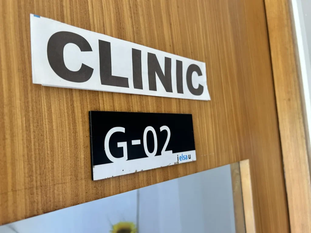필리핀 세부 엘사(ELSA) 어학원 부대시설 클리닉으로, 원목 문에 CLINIC 표지판과 G-02 호실 명판이 붙어 있는 의무실 출입문입니다.