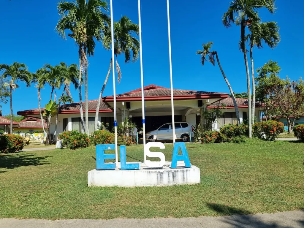필리핀 세부 엘사(ELSA) 어학원 캠퍼스 전경으로, 잔디밭 위 파랑·흰색 입체 'ELSA' 조형물과 국기게양대, 뒤편 본관이 보이는 모습입니다.