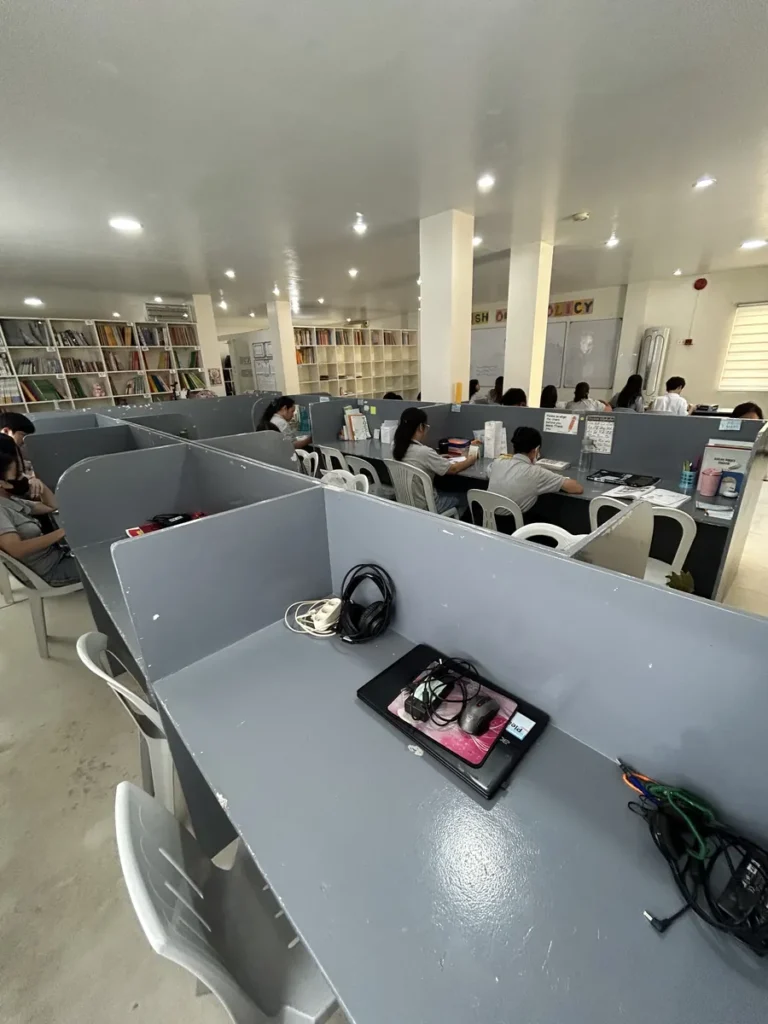 필리핀 세부 JJES 어학원 일대일 강의실로, 칸막이 책상이 여러 줄로 이어지고 앞쪽 책상 위에 헤드폰과 노트북이 놓인 넓은 교실입니다.