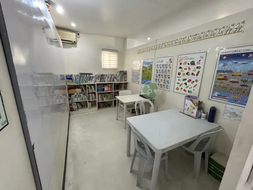 필리핀 세부 JJES 어학원 일대일 강의실로, 동화책이 꽂힌 책장과 흰색 테이블, 에어컨이 있는 작은 학습방 모습입니다.