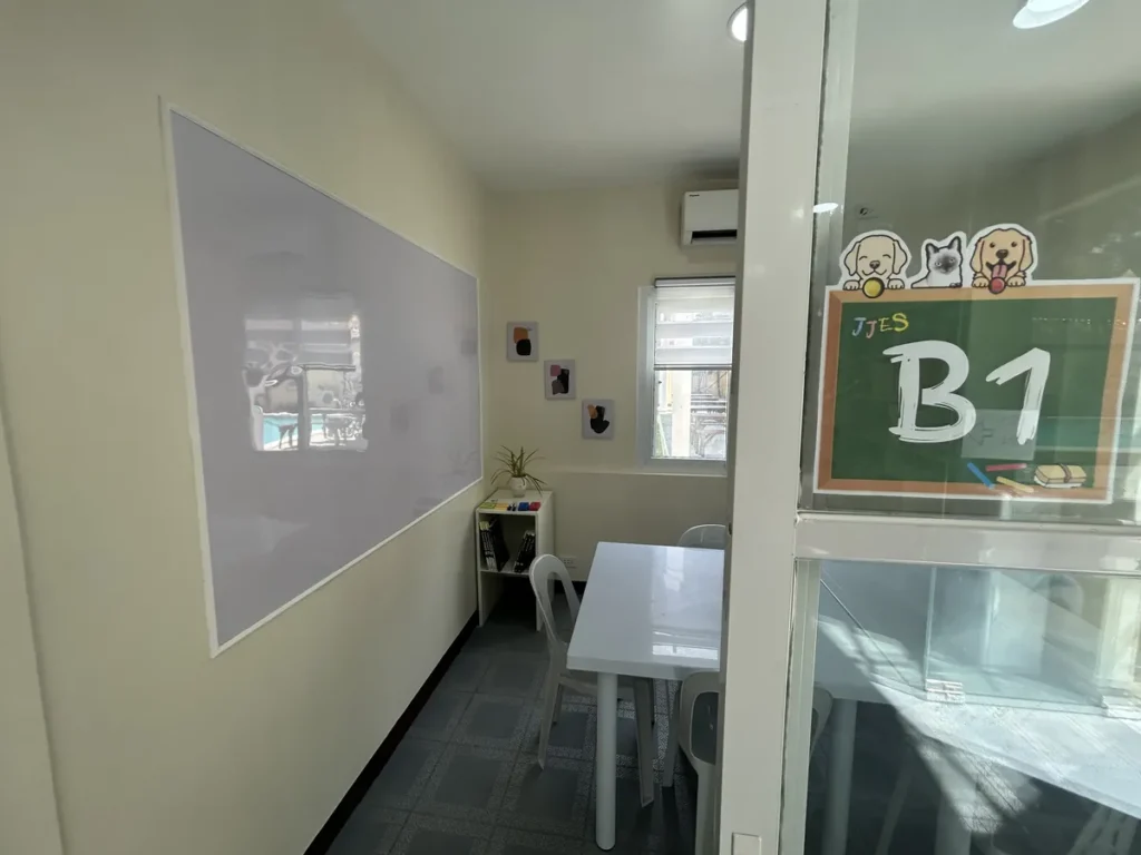 필리핀 세부 JJES 어학원 그룹 강의실로, 복도 안쪽 테이블 너머 유리문에 강아지 그림과 JJES B1 반 팻말이 붙어 있는 모습입니다.