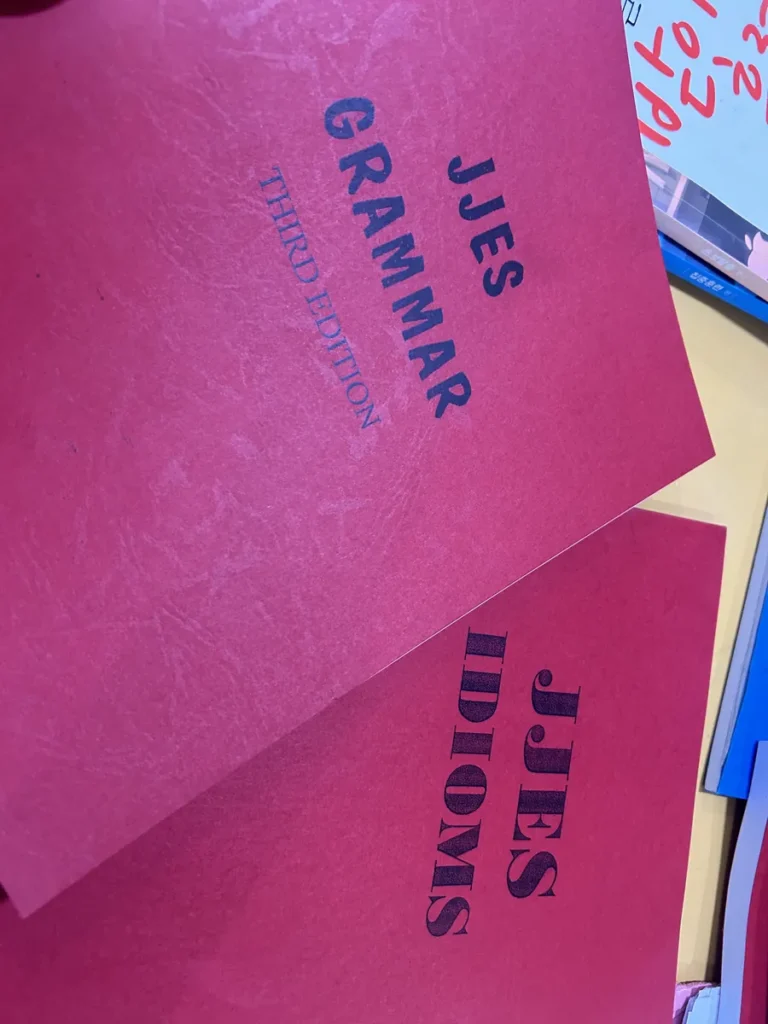 필리핀 세부 JJES 어학원 강의실 교재로, 빨간 표지의 JJES Grammar와 JJES Idioms 교재가 겹쳐 놓인 모습입니다.