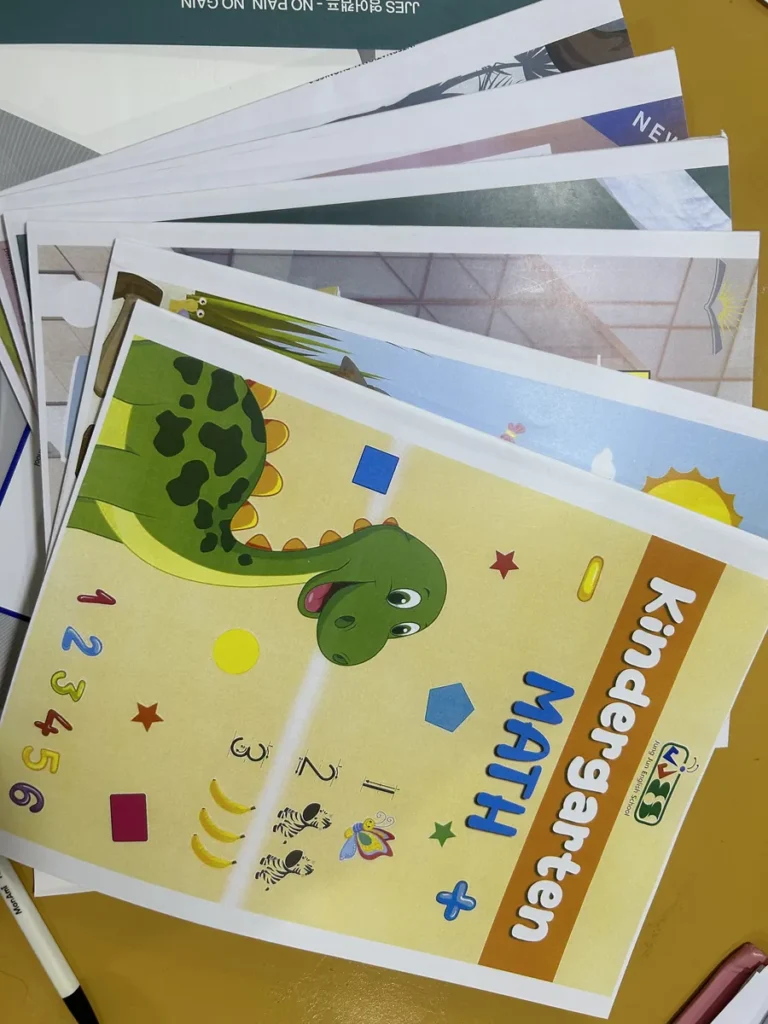필리핀 세부 JJES 어학원 강의실 교재로, 공룡 그림의 Kindergarten Math 교재가 맨 위에 놓이고 여러 교재가 부채꼴로 펼쳐진 모습입니다.