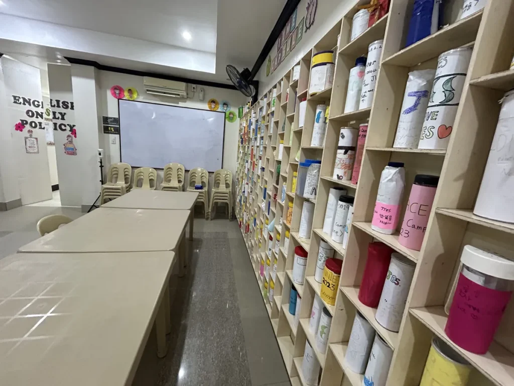 필리핀 세부 JJES 어학원 도서관 내부로, ENGLISH ONLY POLICY 안내판과 화이트보드 옆으로 칸막이 수납장이 길게 이어지는 모습입니다.