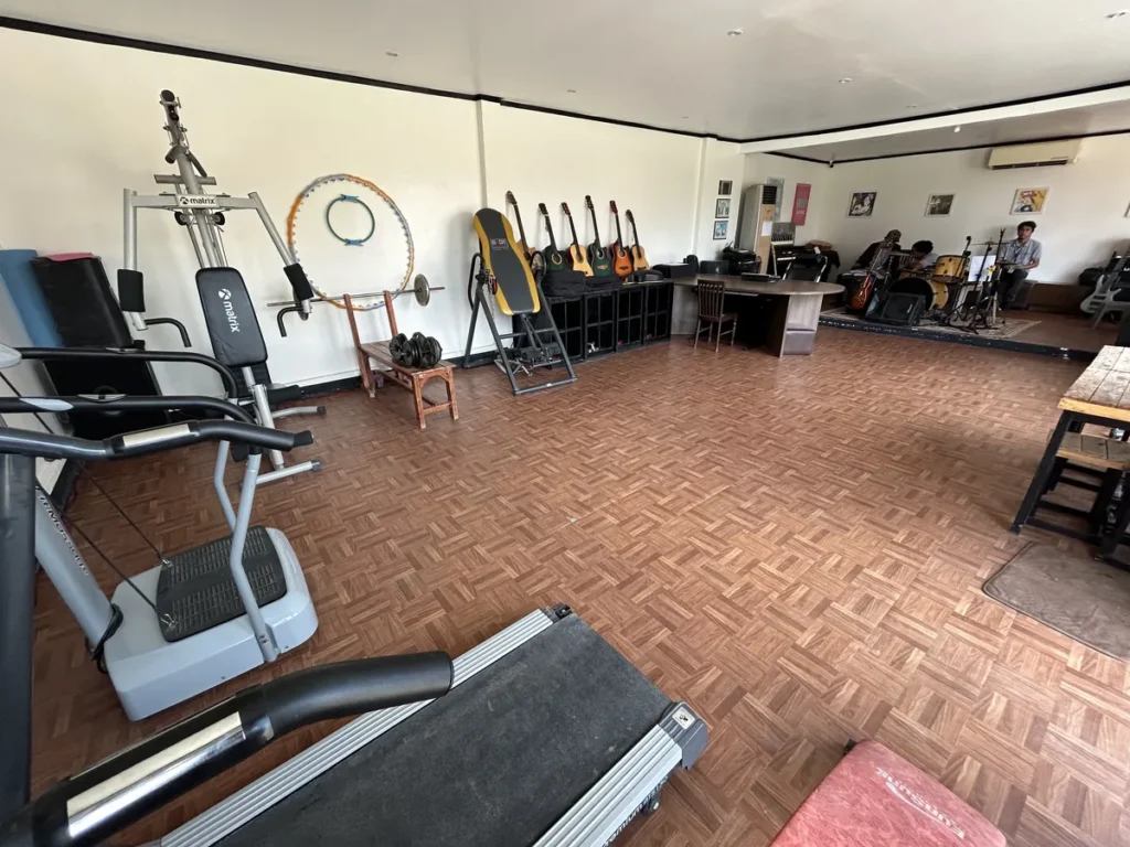 필리핀 세부 JJES 어학원 부대시설 음악실&헬스장으로, 러닝머신과 근력기구 뒤로 기타가 걸린 벽과 무대 쪽 밴드 연습 모습이 함께 보이는 전경입니다.