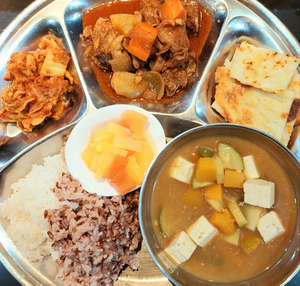 필리핀 세부 JJES 어학원 학생식당 식사로, 매콤한 닭볶음탕과 구운 빵·과일에 밥과 두부 된장찌개를 곁들인 식판입니다.