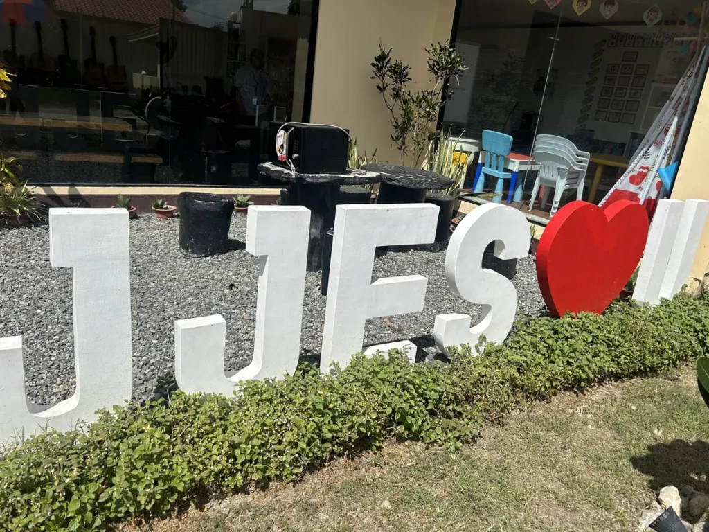 필리핀 세부 JJES 어학원 학원전경 외관으로, 통유리창 안으로 기타와 어린이 의자가 비치는 건물 앞 자갈마당의 JJES 입체 글자와 하트 조형물입니다.