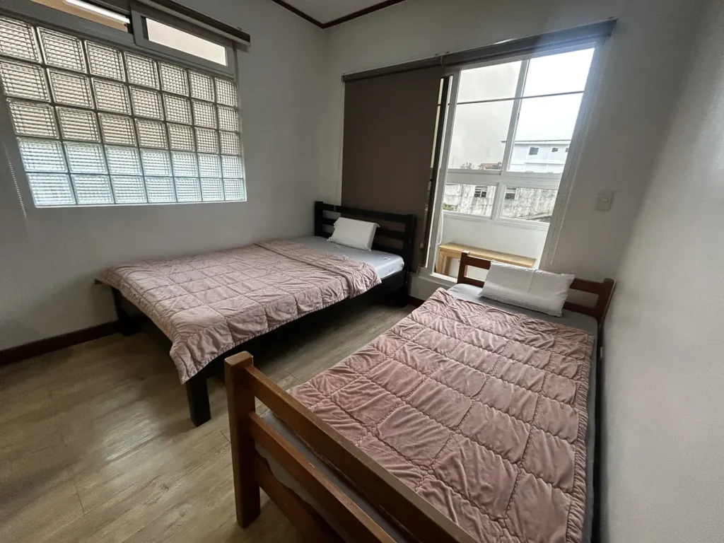 필리핀 바기오 웨일즈(WALES) 어학원 기숙사 P스튜디오 침실로, 핑크 퀼트 이불의 침대 두 개와 유리블록 창, 작은 발코니가 함께 있는 공간입니다.