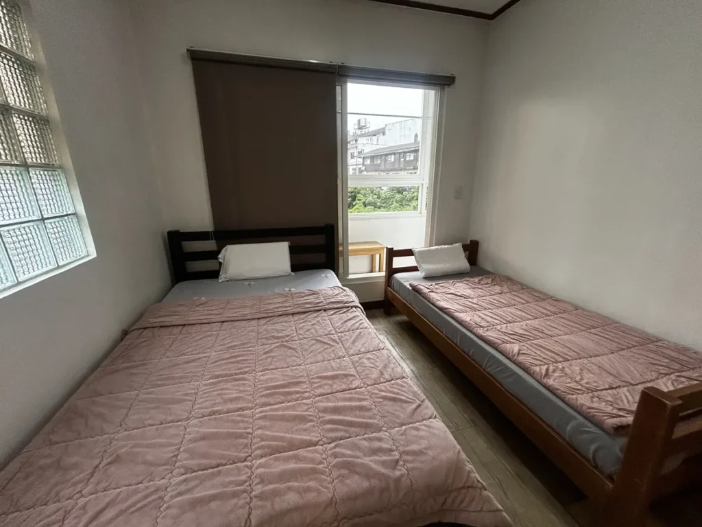 필리핀 바기오 웨일즈(WALES) 어학원 기숙사 P스튜디오 침실로, 핑크 퀼트 침구의 침대 두 개와 창밖으로 시내 건물과 초록 나무가 보이는 공간입니다.