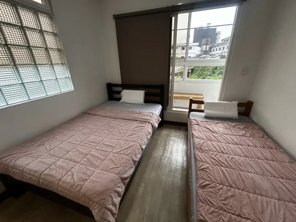 필리핀 바기오 웨일즈(WALES) 어학원 기숙사 P스튜디오 침실로, 핑크 퀼트 침대 두 개와 유리블록 창, 발코니 창이 보이는 넓은 침실입니다.
