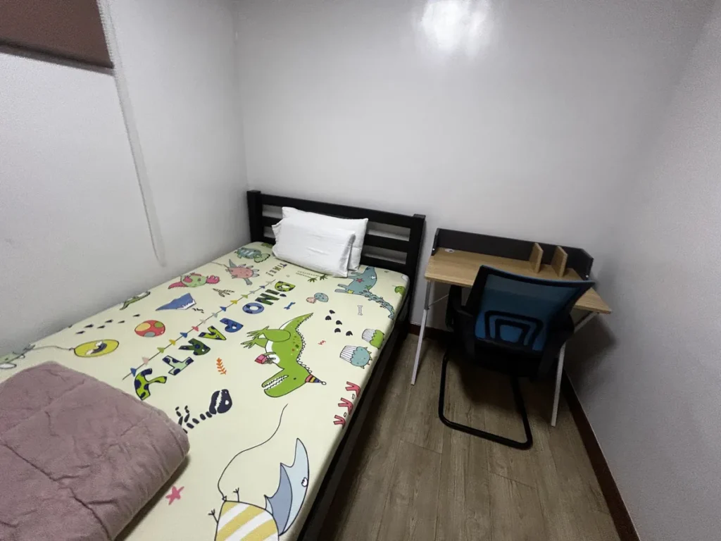 필리핀 바기오 웨일즈(WALES) 어학원 기숙사 1인실 공유형으로, 공룡 무늬 이불과 베개가 정돈된 싱글 침대 옆에 책상·의자가 놓인 모습입니다.