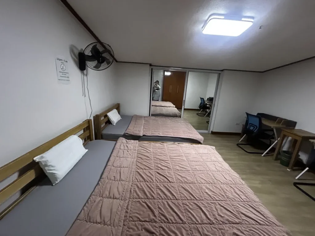 필리핀 바기오 웨일즈(WALES) 어학원 기숙사 2인실 공유형으로, 핑크빛 침구 침대 두 개와 거울에 비친 책상, 의자가 담긴 객실입니다.