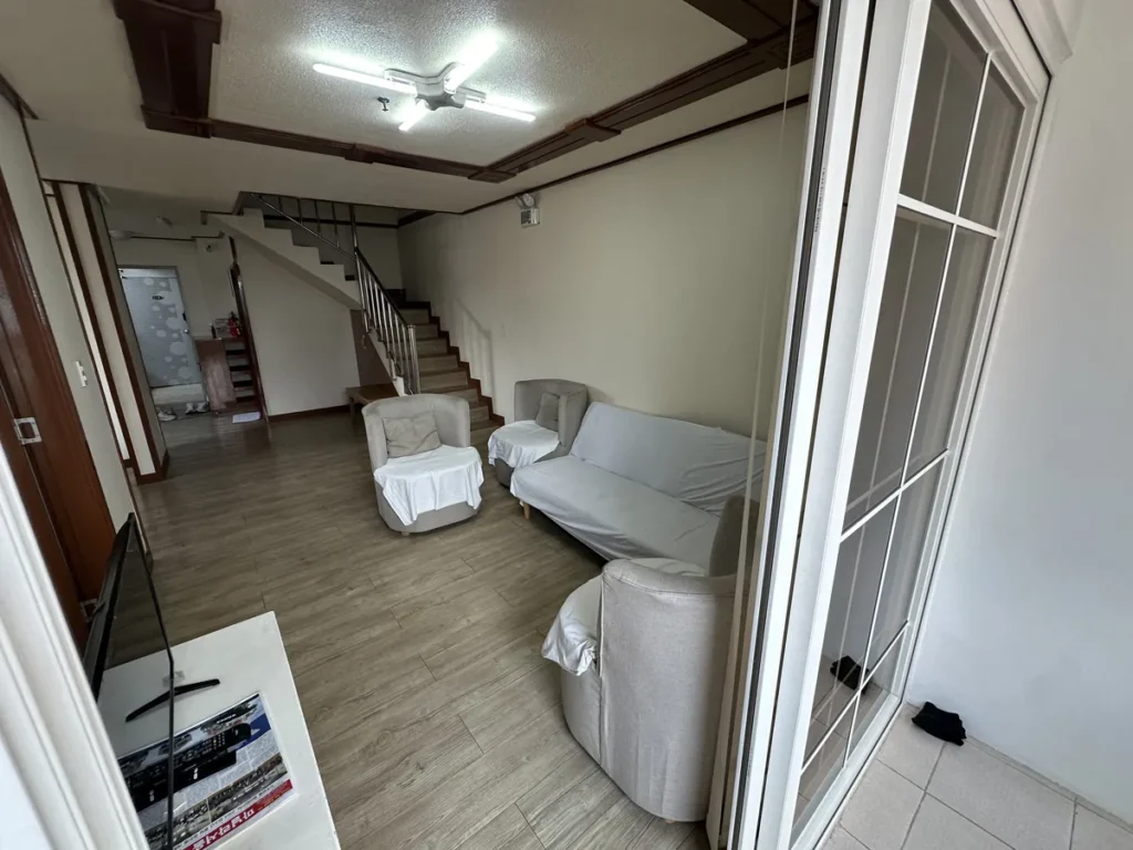 필리핀 바기오 웨일즈(WALES) 어학원 기숙사 공유형 공용공간 거실로, 흰 커버 씌운 연회색 소파와 안락의자, 2층 계단이 보이는 공간입니다.