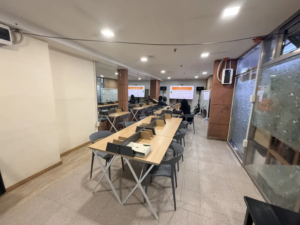필리핀 바기오 웨일즈(WALES) 어학원 그룹강의실로, 칸막이가 달린 책상이 줄지어 놓인 학습홀에 주황색 발표 화면이 벽면 TV에 띄워진 모습입니다.