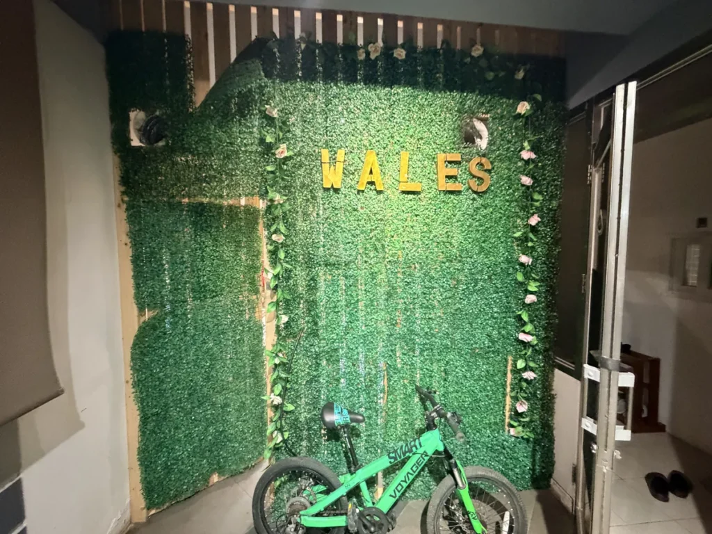 필리핀 바기오 웨일즈(WALES) 어학원 부대시설 휴게공간으로, 초록 이끼 월에 금색 WALES 글자와 자전거가 있는 포토존입니다.