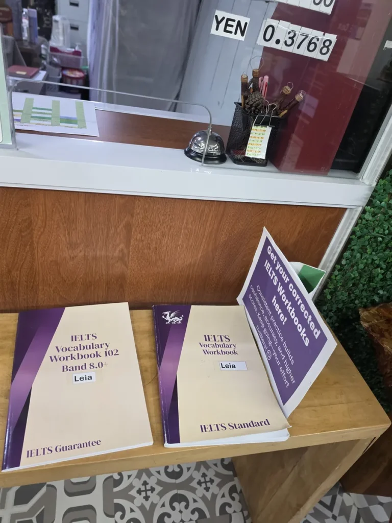 필리핀 바기오 웨일즈(WALES) 어학원 부대시설 오피스·안내판으로, 원목 테이블에 IELTS 보카북과 교정 워크북 안내문이 놓인 모습입니다.