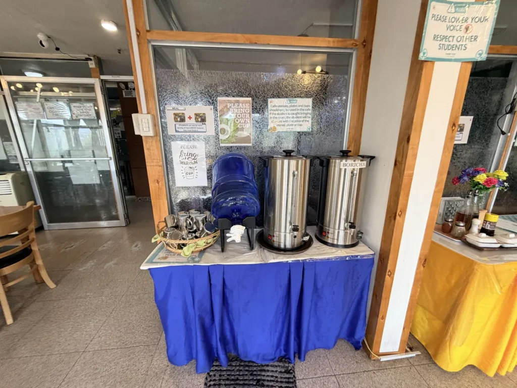 필리핀 바기오 웨일즈(WALES) 어학원 식당으로, 파란 테이블보 위 보리차·생수 디스펜서와 정숙 안내판이 함께 놓인 셀프 음료대 모습입니다.