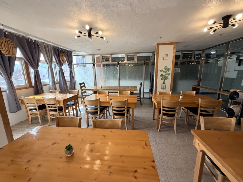 필리핀 바기오 웨일즈(WALES) 어학원 식당으로, 라탄 조명 아래 원목 테이블이 늘어서 있고 안쪽 유리 파티션 너머로 공간이 이어지는 모습입니다.