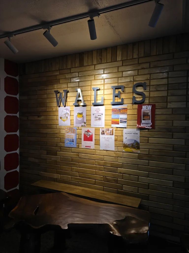 필리핀 바기오 웨일즈(WALES) 어학원 학원전경 입구로비로, 나무 벽돌 벽에 걸린 WALES 로고와 안내 포스터, 원목 벤치를 가까이서 담은 모습입니다.