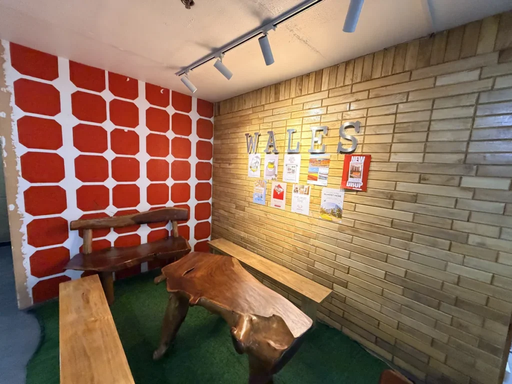 필리핀 바기오 웨일즈(WALES) 어학원 학원전경 입구로비로, WALES 글자 사인 아래 원목 벤치와 테이블, 인조잔디 바닥을 갖춘 라운지 공간입니다.