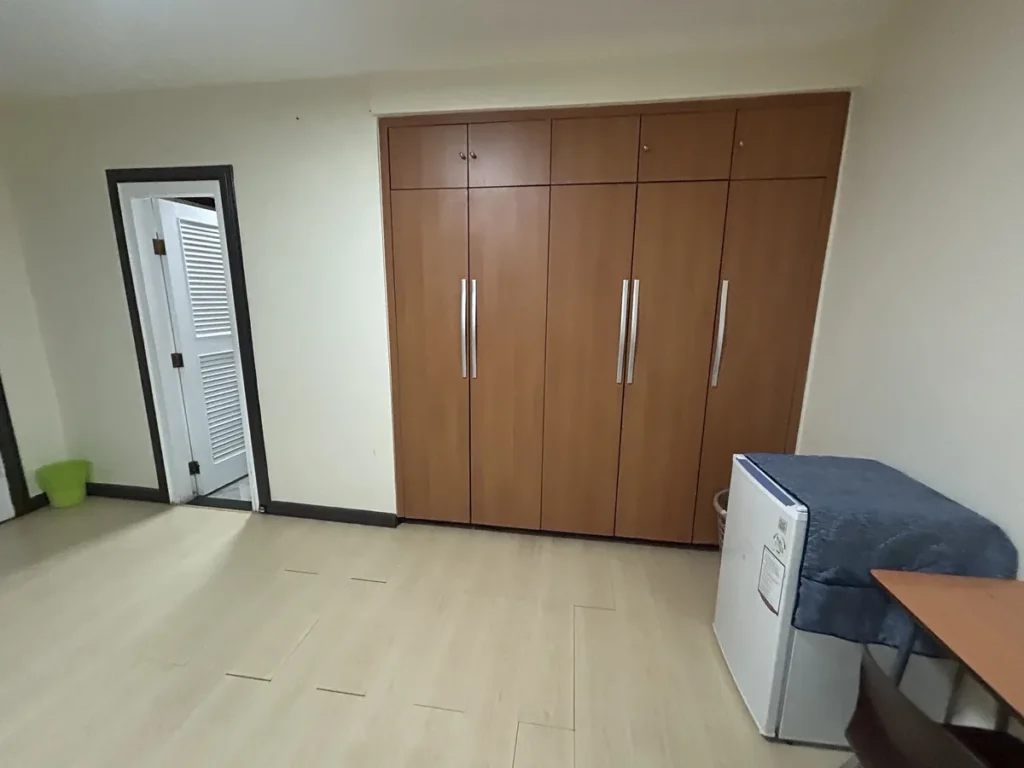 필리핀 세부 CIJ 어학원 3인실로, 원목 붙박이 옷장과 덮개를 씌운 미니 냉장고, 열린 화장실 문이 함께 보이는 객실 한쪽 모습입니다.