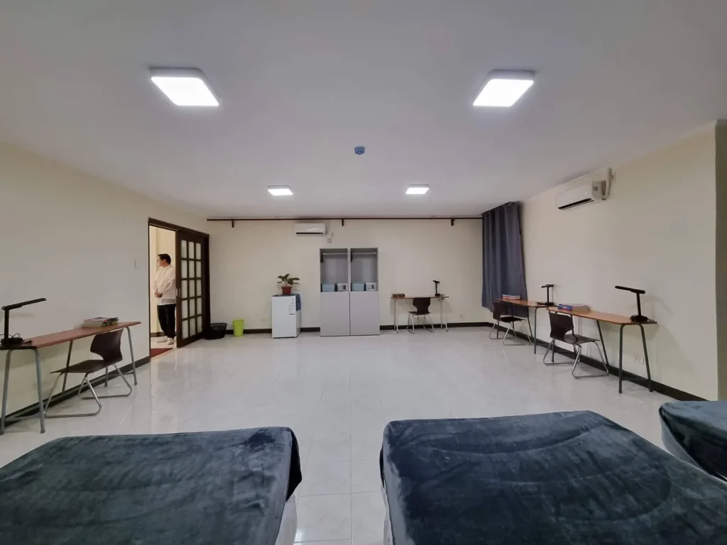 필리핀 세부 CIJ 어학원 4~6인실로, 침대 쪽에서 바라본 맞은편에 개인 책상과 의자·옷장·소형 냉장고·벽걸이 에어컨이 놓인 모습입니다.