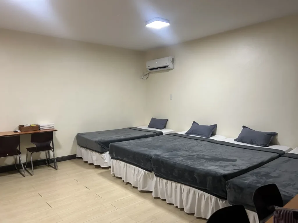 필리핀 세부 CIJ 어학원 4~6인실로, 밝은 원목 무늬 바닥에 회색 담요 침대 세 개가 놓이고 벽걸이 에어컨과 책상이 있는 아늑한 방입니다.