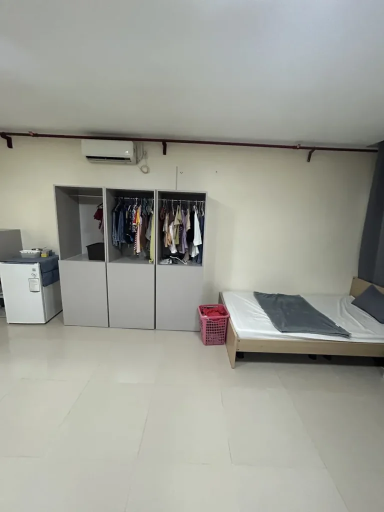 필리핀 세부 CIJ 어학원 4~6인실로, 옷이 걸린 오픈형 옷장 세 칸과 소형 냉장고·빨래 바구니·원목 프레임 싱글 침대가 놓인 벽면입니다.