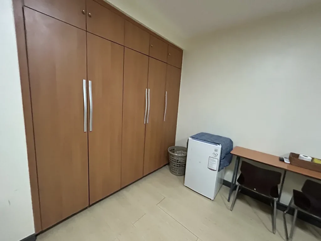 필리핀 세부 CIJ 어학원 4~6인실로, 벽면 전체를 채운 원목 붙박이 옷장과 소형 냉장고·빨래 바구니, 의자 두 개가 놓인 책상이 있는 모습입니다.