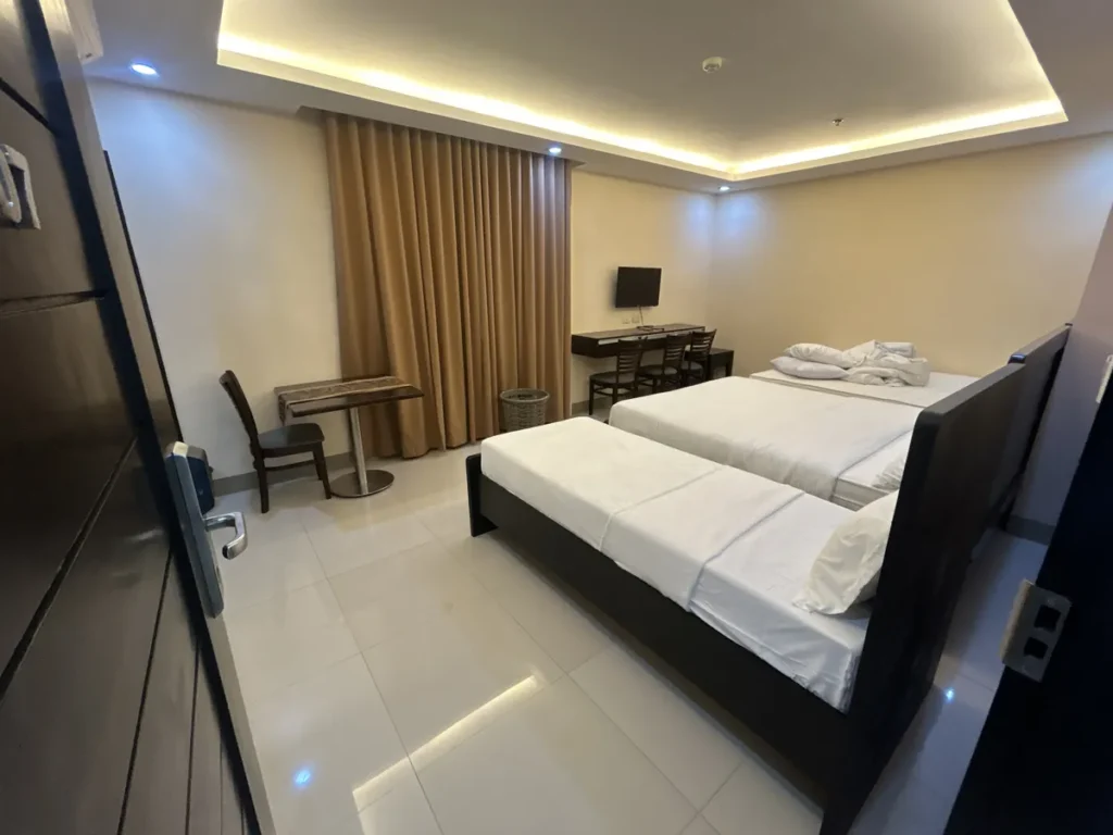 필리핀 세부 CIJ 어학원 외부 기숙사 3인실로, 출입문에서 바라본 간접조명 천장 아래 침대 3개와 책상·식탁이 놓인 넓은 객실 전경입니다.