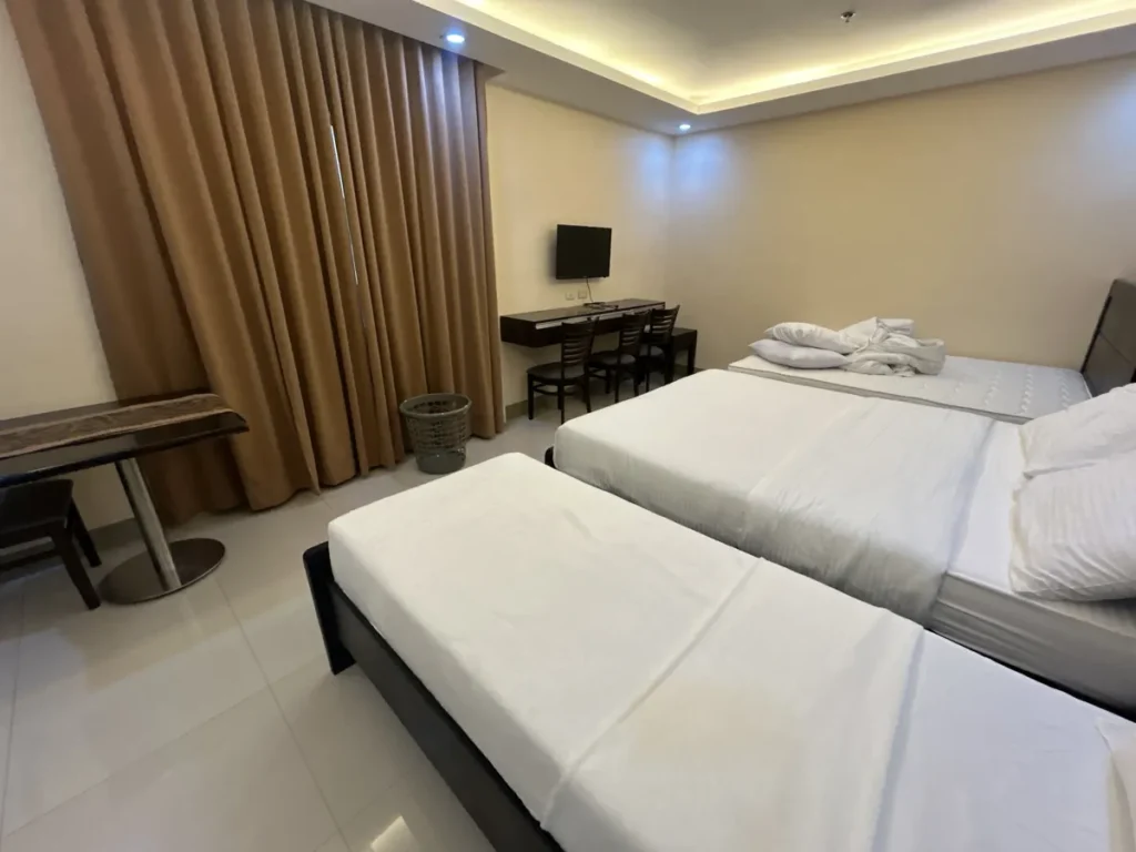 필리핀 세부 CIJ 어학원 외부 기숙사 3인실로, 흰 침구의 침대 3개가 나란히 놓이고 커튼 옆으로 TV와 3인용 책상이 보이는 객실입니다.
